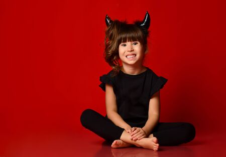 Little brunette girl with devil horns, ponytail, in black blouse and leggings, barefoot. She is smiling, sitting on floor in lotus position against a red background. Masquerade, Halloween. Close upの写真素材