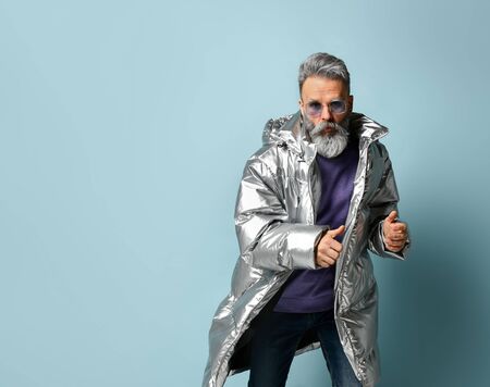 Gray-haired, bearded, mature man in purple pullover and sunglasses, silver colored down jacket, jeans. He is running, showing thumb up, posing on blue background. Fashion, style. Close up, copy spaceの写真素材