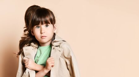 a little brunette girl is holding a raincoat with her arms crossed over her chest. In a green blouse and blue jeans. On a beige background in the Studioの写真素材