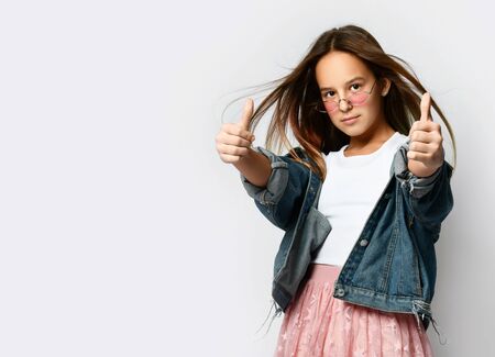 Young teen girl in stylish casual clothes and pink sunglasses standing and showing good thumb sign...