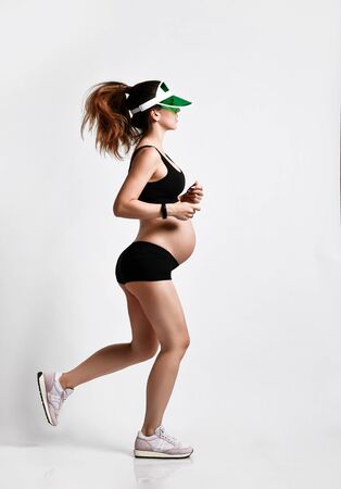 Pregnant girl with belly, in green sun visor, black sport shorts and top, pink sneakers. She is jogging isolated on white background. Family and maternity concepts. Full-length, copy space, side viewの写真素材