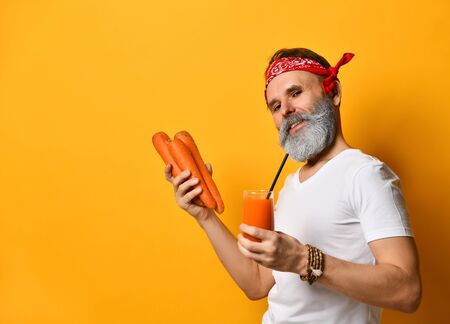 Gray-bearded grandpa in red bandana, white t-shirt, bracelet. He smiling, holding glass of fresh squeezed juice with tube, three big carrots, posing sideways on yellow background. Close up, copy spaceの写真素材