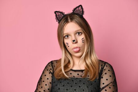 Blonde teenager in black dress, headband in form of cat ears, face painting. She is showing her tongue, posing on pink background. Beauty, fashion, advertising. Copy space. Close upの写真素材