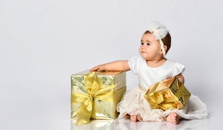 Little kid in dress and headband, barefoot. She holding two golden gift boxes with bows, sitting on floor isolated on white. Christmas, New Year, birthday. Childhood, advertising for babies. Close upの写真素材