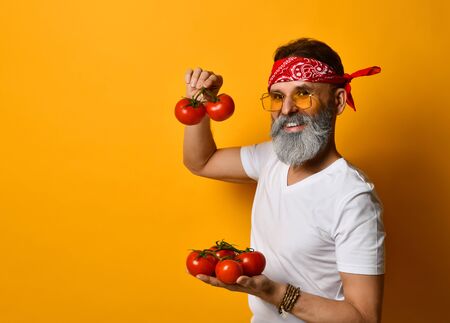 Gray-bearded grandpa in red bandana, white t-shirt, sunglasses and bracelet. He smiling, showing red tomatoes on twigs, posing sideways against orange background. Close up, copy spaceの写真素材
