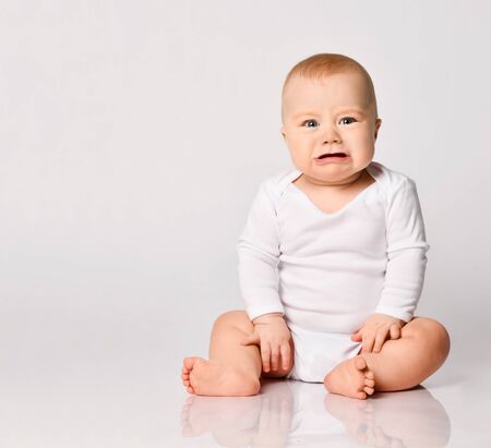 Chubby little kid in bodysuit, barefoot. He is about to cry, sitting on floor isolated on white studio background. Concept for articles about childhood or advertising for babies. Close up, copy spaceの写真素材