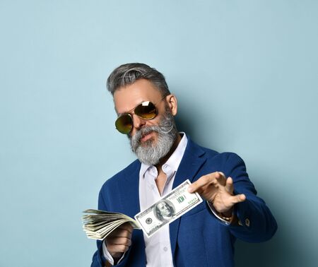 Boss male in white shirt, jacket and sunglasses. He is smiling, holding some hundred dollar bills and offering you to take one, posing on blue studio background. Close up, copy spaceの写真素材