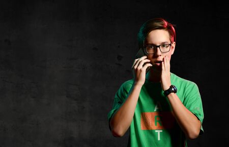 Young concentrated teen boy in green t-shirt and glasses standing, talking on phone and looking...