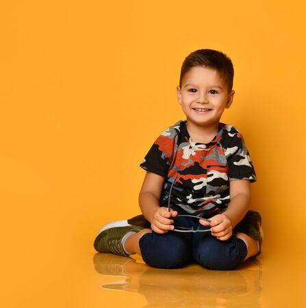 Little brunet child in camouflage t-shirt, blue shorts and khaki sneakers. He smiling, sitting on floor, holding gray sunglasses, posing on orange background. Childhood, fashion. Close up, copy spaceの写真素材