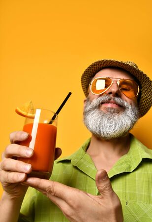 Gray-bearded man in a hat, green shirt, sunglasses at the resort. He smiles and gives a glass with a cocktail with a slice of lemon and a tube, posing against an orange background. Close copy spaceの写真素材