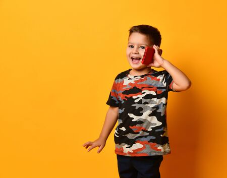 Little brunet boy in camouflage t-shirt is smiling, talking by red smartphone, looking at you, posing against orange studio background. Happy childhood, gadget, advertising. Close up, copy spaceの写真素材