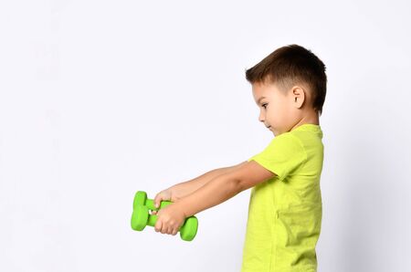 Little male in yellow t-shirt and denim shorts. He smiling, raising his hand up holding green dumbbell, posing isolated on white studio background. Concept of childhood and sport. Close up, copy spaceの写真素材