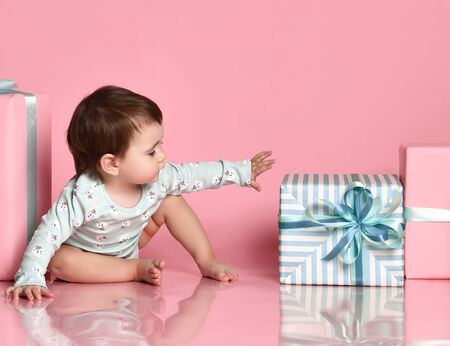 The baby in the bodysuit reaches for a gift box, beautifully wrapped for her mother. on a pink background. Women's Day Celebration. profile faces side viewの写真素材