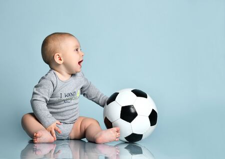 Child in gray bodysuit, barefoot. He holding soccer ball, tongue out, sitting on floor against blue background. Concept for articles about childhood or advertising for babies. Close up, copy spaceの写真素材