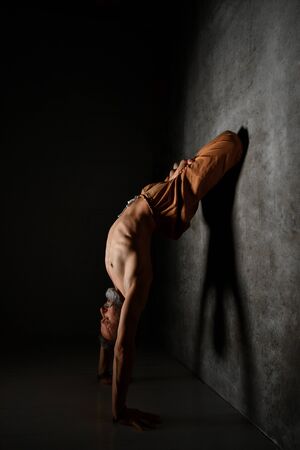 Gray-haired, bearded yogi male with naked torso, in brown pants is practicing yoga. Standing on his hands, leaning against a wall by legs being in lotus pose. Flexible and plastic. Twilight. Close upの写真素材