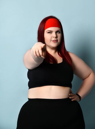 Hey, go on a diet! Fat redhead girl in a red headband, black top. She points at you, posing on a blue studio background. Positive body, sports concept.の写真素材