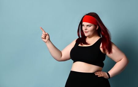 Overweight redhead woman in red headband, black top and leggings, fitness bracelet. She pointing at...