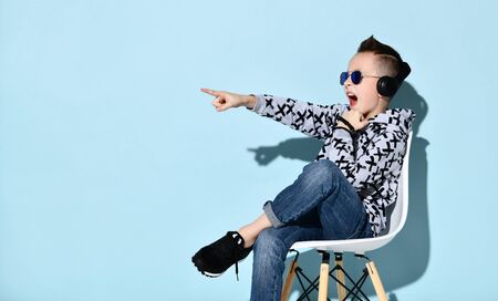 Teen boy in headphones and sunglasses, blue jeans, stylish hoodie, black sneakers. Pointing at something, sitting on white chair against blue background. Childhood, fashion. Close up, copy spaceの写真素材