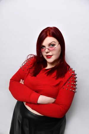 Overweight red-haired woman in a red studded top, black leather skirt and earrings. She stands with her arms folded in a twisted pose.の写真素材