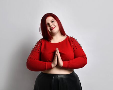 Overweight red-haired woman in a red studded top, black leather skirt and earrings. She folded her palms and begged you while posing isolated on whiteの写真素材