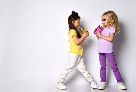 Two little gorgeous girls in sunglasses, colorful casual clothes. They holding cocktail bottles, smiling, looking at each other, posing isolated on white. Childhood, fashion. Full length, copy spaceの写真素材