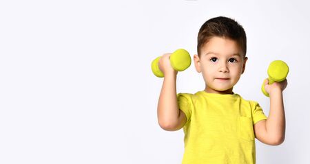 Little male in yellow t-shirt and denim shorts. He smiling, raising his hand up holding yellow dumbbell, posing isolated on white studio background. Concept of childhood and sport. Close up, copy spaceの写真素材