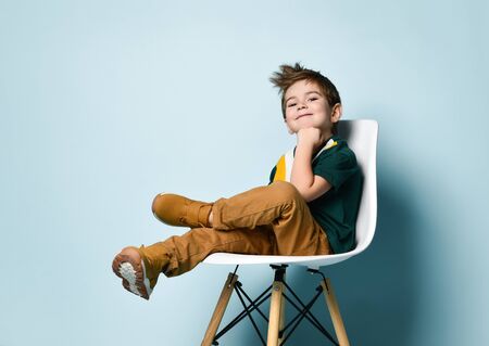 Little brunet kid in colorful t-shirt, brown pants and sneakers. Smiling, propping his chin by fist, sitting on white chair against blue studio background. Childhood, fashion. Close up, copy spaceの写真素材