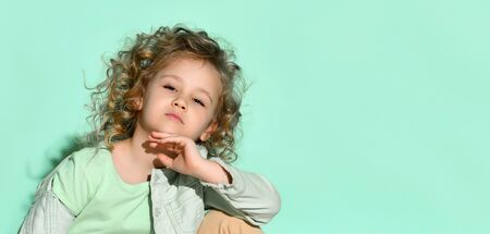 Little blonde curly girl in t-shirt, denim jacket and beige pants. She propped her chin by hand,...