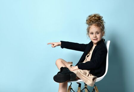 Blonde little model with hairstyle, in beige dress, black jacket and boots. She pointing at something, sitting cross-legged on white chair against blue background. Close up, copy space, side viewの写真素材