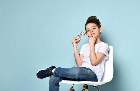 Concentrated schoolboy sits on chair holding dollars and touching chin with hand thinking how to...