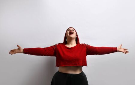 Fat ginger female in red spiked top, black leather skirt and earrings. She has spread her arms and screaming, posing isolated on white photo background. Fashion and style. Close up, copy spaceの写真素材