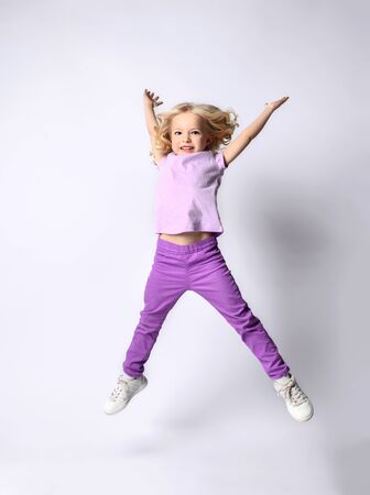Little blonde curly kid dressed in purple blouse and pants, white sneakers. She is smiling while jumping up with raised hands, posing isolated on white. Childhood, fashion. Full length, copy spaceの写真素材