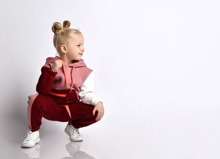 Little blonde kid with buns hairstyle, in colorful tracksuit, sneakers. She smiling, squatting, showing fist, posing isolated on white. Childhood, fashion, advertising and sport. Close up, copy spaceの写真素材