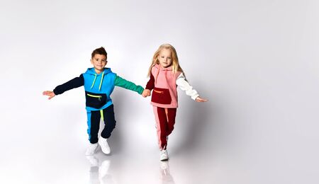Children, boy and girl, in colorful sport suits and sneakers. They are holding hands, running, isolated on white studio background. Childhood, fashion, advertising.の写真素材