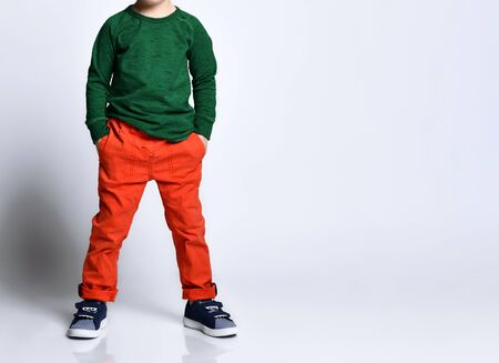 Cropped photo of a boy in a green jumper, orange pants, blue sneakers. He put his hands in his pockets, posing isolated on a white background. Childhood, fashion, advertising. Full length, copy spaceの写真素材