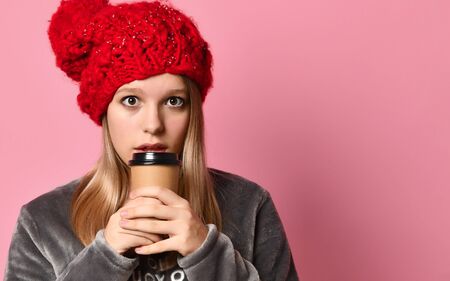 Blonde teenage girl in gray plush pajamas and red hat with pompom. She is holding a paper cup and...