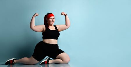 Plus size redhead woman in red headband, black top, shorts, sneakers, fitness bracelet. Showing muscles, posing standing on knees against blue background. Body positive, sport.の写真素材