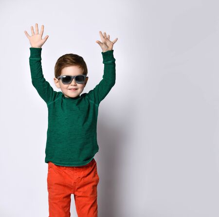 Cute boy in sunglasses, green jumper and orange pants. He raised his hands up and smiling, posing isolated on white studio background. Childhood, fashion, advertising. Close up, copy spaceの写真素材