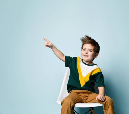 Little brunet kid in colorful t-shirt and brown pants. Smiling and pointing at something by forefinger, sitting on white chair against blue studio background. Childhood, fashion. Close up, copy spaceの写真素材