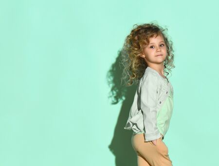 Curly baby in a T-shirt, denim jacket, beige pants. She is smiling standing sideways on a turquoise background. Childhood, fashion.の写真素材