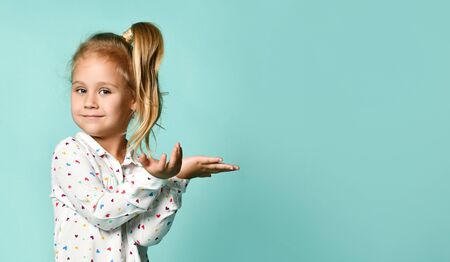 awesome girl with ponytail, dressed in shirt with hearts print. She smiling, acting like holding something in her hands, posing on blue background. Childhood, fashion, advertising. Close up, copy spaceの写真素材