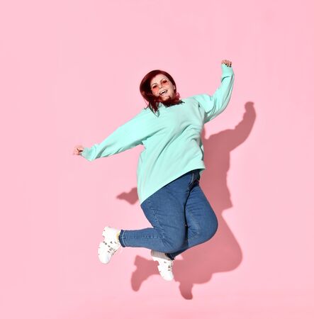 Active young female with overweight in casual attire and glasses jumping up full of happiness and joy. Self esteem, active way of life, charming plus-size, body positive. Studio shot isolated on pinkの写真素材