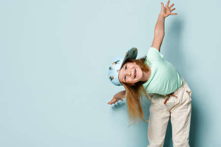 Lovely blonde kid in t-shirt, white pants and cap printed by pineapples. She leaned forward with her arms wide apart and smiling, posing against turquoise studio background. Close up, copy spaceの写真素材