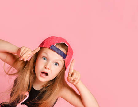 Little blonde female in cap and black blouse. She pointing up by forefingers and looking surprised while posing against pink studio background. Childhood, emotions, advertising. Close up, copy spaceの写真素材