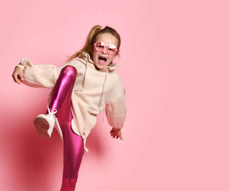 expressive little girl poses on one leg, raising her knee and spreading her arms apart to find balance. Full length shot isolated on pink. Fashion for children, facial expression, games and entertainment.の写真素材