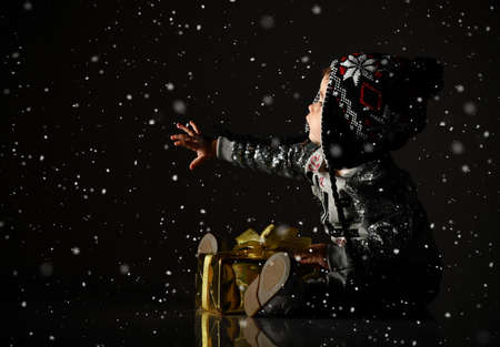 Baby in gray hat, sparkling suit and boots. She looking at snowflakes, posing with golden gift box, sitting on floor against black studio background. Christmas, New Year, holidays. Snowing itの写真素材