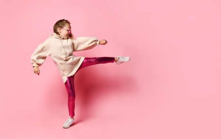 Side view of a little girl in a pink sweatshirt with a hood, shiny leggings and glasses, standing on...