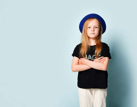 Cute blonde child in blue hat, black t-shirt and white pants. She is looking at you with crossed hands while posing against turquoise studio background. Childhood, fashion, advertising. Copy spaceの写真素材