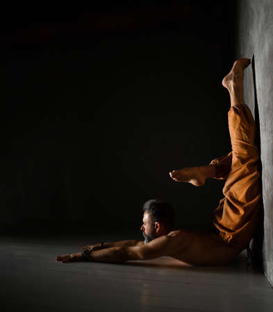 Gray-haired, barefoot, elderly yogi male with naked torso, in brown pants and bracelets is practicing yoga. Lying on the floor, leaning against wall by his legs. Flexible, plastic. Twilight. Close upの写真素材