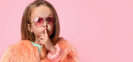Blonde little female in sunglasses, turquoise t-shirt and faux fur coat. She is showing be quiet sign by forefinger while posing on pink background. Happy childhood, gestures. Close up, copy spaceの写真素材